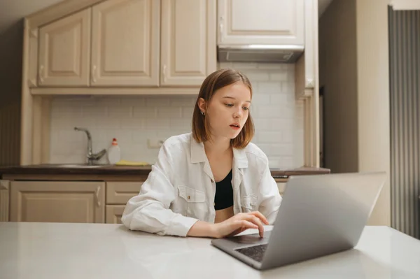 Retrato de uma estudante sentada em uma mesa na cozinha e usando um laptop, com um rosto sério olhando para a tela e estudando.Mulher bonita trabalhando no laptop à mesa em casa no apartamento — Fotografia de Stock