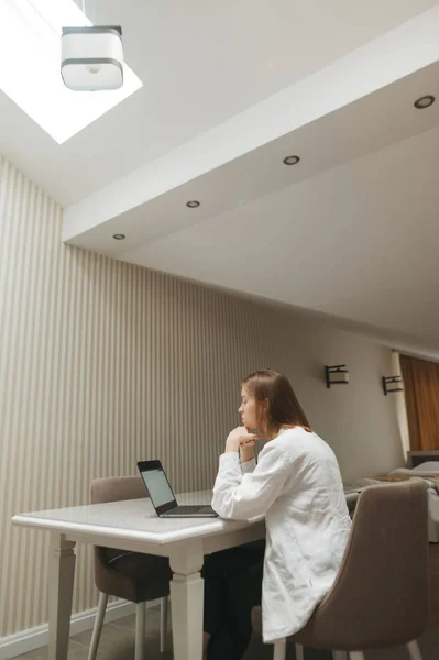 Menina freelancer atraente trabalhando no laptop na mesa no apartamento sótão em estilo escandinavo. Mulher em roupas leves senta-se na sala do sótão na cozinha à mesa com laptop, olha para a tela. Vertical — Fotografia de Stock
