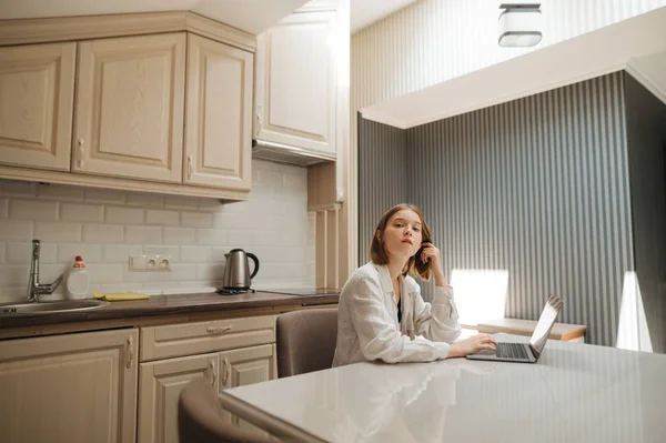 Πορτρέτο ενός χαριτωμένου κοριτσιού που φοράει λευκό πουκάμισο, κάθεται σε ένα τραπέζι στην κουζίνα, χρησιμοποιώντας ένα φορητό υπολογιστή και κοιτάζοντας την κάμερα. Κορίτσι ελεύθερος επαγγελματίας εργάζεται στο σπίτι σε διαμέρισμα στο φορητό υπολογιστή. — Φωτογραφία Αρχείου