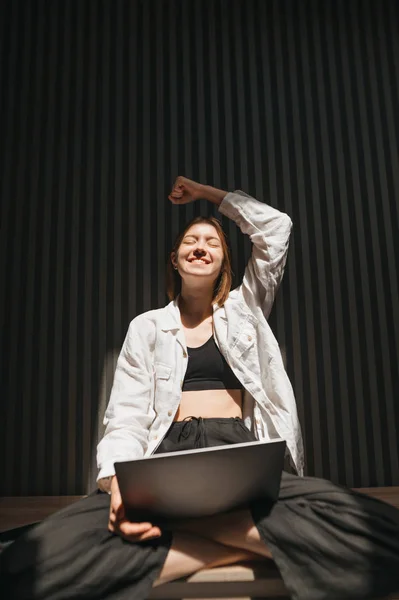 Glad flicka med laptop i knät sitter på bänken mot mörk vägg bakgrund och glädjer. I rummet, ett porträtt av en glad frilansande flicka vann, räckte upp handen. — Stockfoto