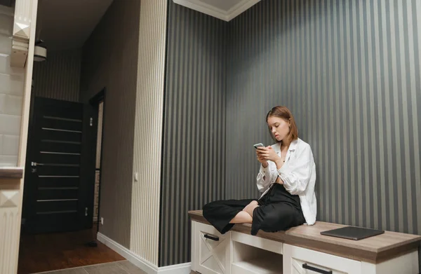 Красива дівчина в стильному одязі сидить вдома на тумбочці в квартирі і використовує смартфон, носить білу сорочку і чорні штани. Стильна жінка розслабляється вдома в квартирах . — стокове фото