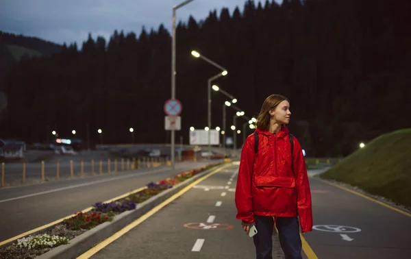 Attractive caucasian girl tourist in a red raincoat looking aside walking on the road in the mountains. Young female traveler enjoying the evening stroll near the mountain forest. — Stock Photo, Image