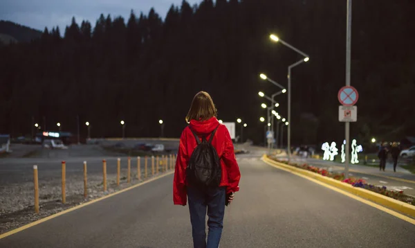 Female traveler in a red raincoat walking down the road in the mountainous scenery in the evening. Young tourist girl back view on the road going to the mountain forest in the street lights. — Stock Photo, Image