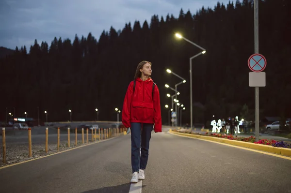 Νεαρή ελκυστική τουρίστρια που περπατάει μόνη της στην εθνική οδό το βράδυ. Γυναίκα ταξιδιώτης σε ένα κόκκινο αδιάβροχο βόλτα κοντά στο βουνό κωνοφόρων δάσος. Ορεινά δάση στο βάθος. — Φωτογραφία Αρχείου