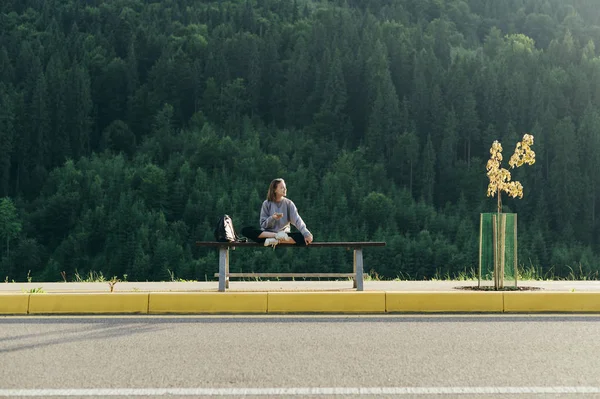 Young attractive female tourist resting on a bench after a forest walk holding smartphone. Positive girl traveler sitting on a bench in a fir forest background. Mountain woods landscape, copy space. — Stock Photo, Image