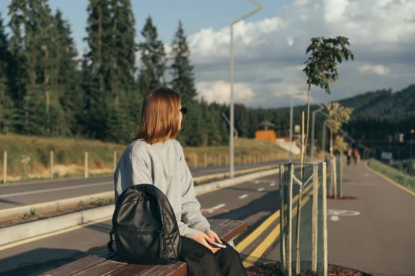 Молода мрійлива жінка-мандрівник сидить на лавці біля дороги між гірськими лісами. Розслаблена туристична дівчина з рюкзаком і сонцезахисними окулярами, що перебувають у горах біля хвойного лісу . — стокове фото