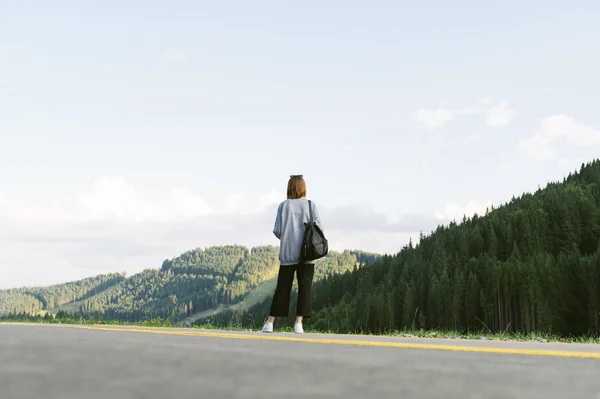Full length photo of a young tourist girl standing on the road looking at beautiful mountain forests scenery. Back view shot of female traveler with backpack enjoying coniferous forests and mountains. — Stock Photo, Image