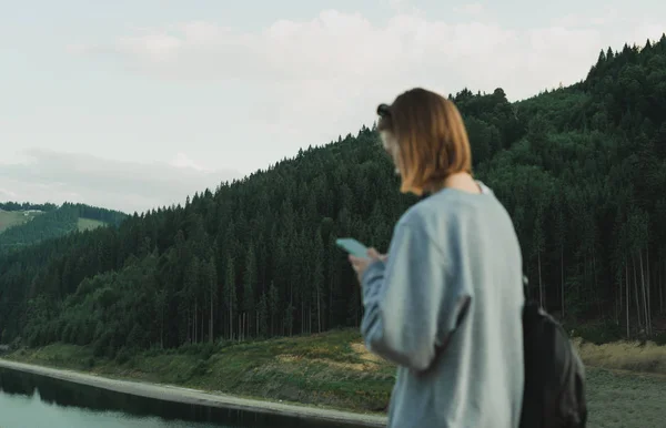 Focus on background. Caucasian female tourist using smartphone app with maps to find the way to coniferous forests in the mountains. Young traveler girl in a grey sweatshirt with backpack in the woods — Stock Photo, Image