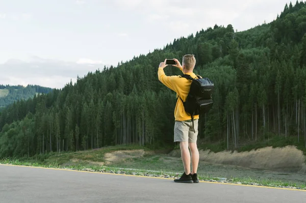 Jovem viajante masculino em camisola amarela tirando foto no smartphone da paisagem da floresta de montanha. Visão traseira do turista usando celular para fotografar madeiras de coníferas. Viagens, conceito de férias . — Fotografia de Stock