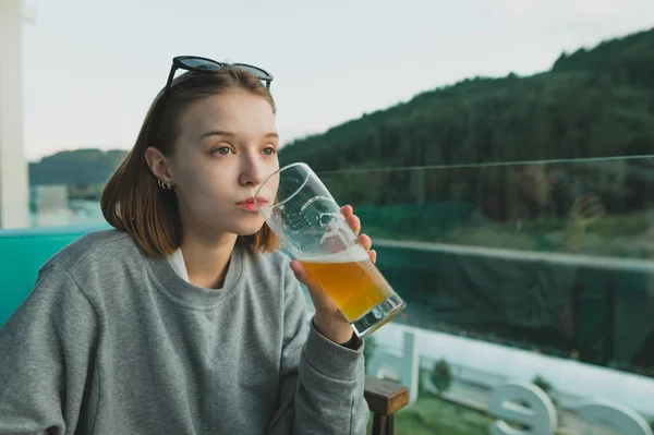 Uma jovem e atenciosa mulher a beber cerveja no bar do terraço, perto da floresta e do lago. Mulher atraente desfrutando de um copo de cerveja em uma paisagem de floresta e lago . — Fotografia de Stock