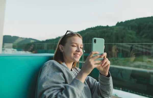 Портрет веселої усміхненої жінки з мобільним телефоном на терасі в гірському лісі та ландшафті озера. Молода жінка з широкою посмішкою фотографує гірські ліси на своєму смартфоні . — стокове фото