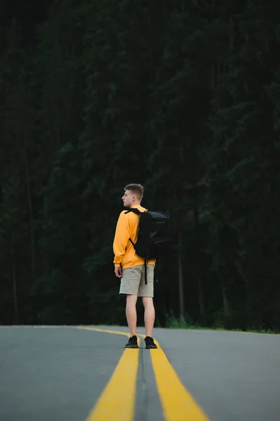 Вид ззаду на чоловічих мандрівників у жовтій сорочці, що стоїть посеред дороги на гірському лісовому ландшафті. Вертикальний постріл туриста з рюкзаком в центрі шосе на лісовому фоні . — стокове фото