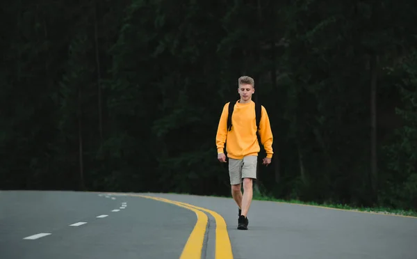 若い男性の旅行者は、山の森の背景にアスファルトの道路を散歩。山の針葉樹林の近くを歩く黄色のスウェットシャツの魅力的な白人観光客. — ストック写真