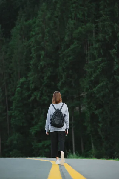 Back view on female traveler in grey sweatshirt standing in the middle of the road at mountain forest landscape. Vertical shot of tourist with backpack in the center of highway on woodland background. — Stock Photo, Image