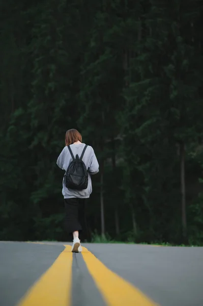 Vertical shot of female traveler in grey sweatshirt and black pants with backpack strolling in the middle of asphalt road in mountain woods. Back view on tourist girl walking near coniferous forest. — Stock Photo, Image