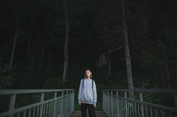 Evening photo of attractive female hiker standing on the bridge near the coniferous woods feeling careless and relaxed. Caucasian tourist girl in the mountain woods, evening walk. Copy space.