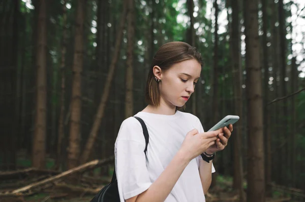 Νεαρή ελκυστική γυναίκα ταξιδιώτης χρησιμοποιώντας το smartphone χάρτες εφαρμογή της για να βρείτε το δρόμο από το ορεινό δάσος. Συγκεντρωμένη τουρίστρια που κρατάει κινητό και στέλνει μηνύματα σε κάποιον ενώ μένει στο δάσος.. — Φωτογραφία Αρχείου
