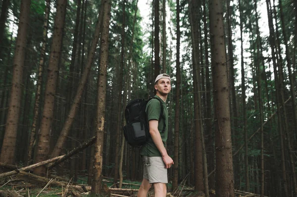 Retrato de un senderista masculino reflexivo con una mochila de pie en el bosque de coníferas de montaña mirando hacia el futuro. Tranquilo, relajado, chico turista descuidado de pie entre abetos en el fondo del bosque . — Foto de Stock