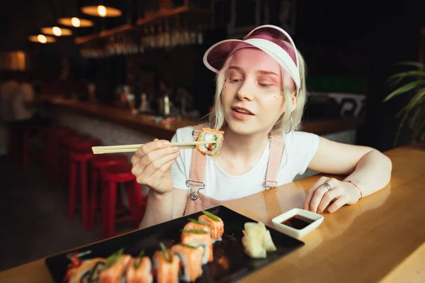 Portret pięknej dziewczyny w różowe ubrania i z makijażu siedzi w azjatyckiej restauracji trzyma laski i będzie jeść Sushi. Kobieta trzyma Sushi Roll na patyku i szuka apetytu. Kuchnia Japońska — Zdjęcie stockowe