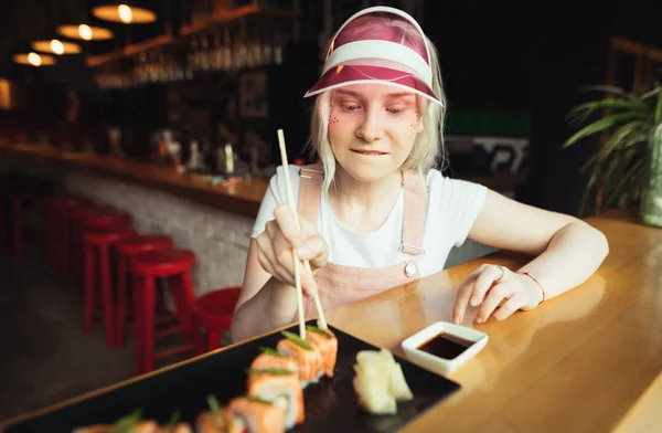 Głodna dziewczyna z pałeczkami w dłoniach bierze rolkę Sushi z jej talerza i szuka jedzenia z apetytem. Teen Girl w różowe ubrania jedzenia ulubionego Sushi w restauracji. Koncepcja kuchni japońskiej — Zdjęcie stockowe