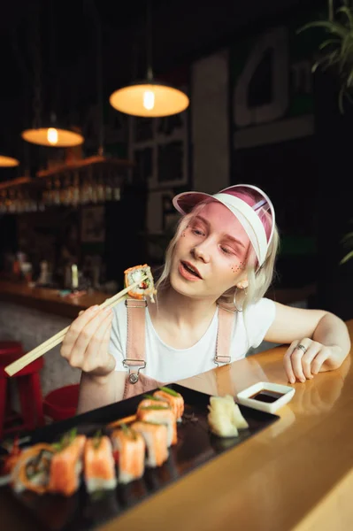 Hübsches Mädchen in pinkfarbener Mütze und niedlicher Kleidung hält eine Rolle Sushi auf Stöcken und steht kurz davor zu essen. Mädchen isst gerne Sushi in einem asiatischen Restaurant mit Appetit auf die Rolle in der Hand. — Stockfoto