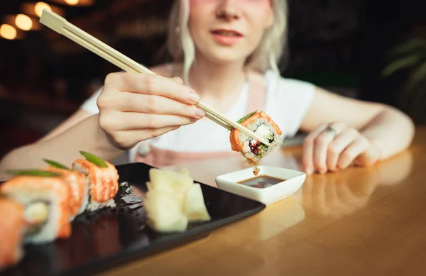 Фон. Крупный план суши в палочках для еды над тарелкой с соевым соусом, девушка сидит за столом в японском ресторане и ест суши-роллы. Женщина погружает суши-ролл в соевый соус . — стоковое фото