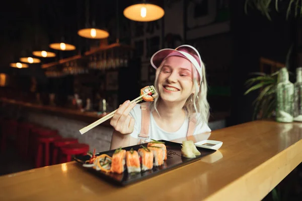 Happy Lady trzyma Sushi Roll na pałeczki, patrzy na pyszne japońskie jedzenie i uśmiechy, siedzi w przytulnej restauracji azjatyckiej. Uśmiechnięta dziewczyna jedzenia sushi na stole w restauracji, ubrany cute różowe ubrania — Zdjęcie stockowe