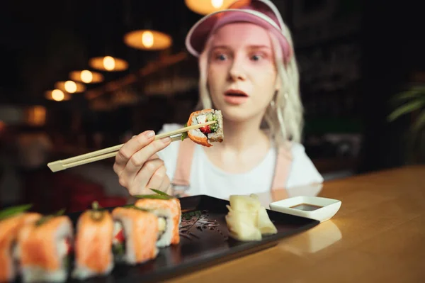 Überraschte Dame mit pinkfarbener Mütze, die Essstäbchen mit Sushi-Rolle in der Hand hält und überrascht auf das Essen blickt. junges Mädchen mit einem schönen Stück Sushi auf Essstäbchen, Nahaufnahme Foto. Hintergrund — Stockfoto