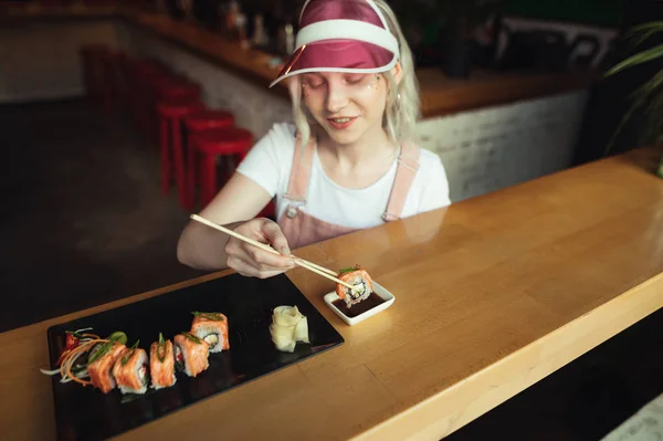 Lächelndes Mädchen legt Sushi-Rolle in Sojasauce, isst Sushi in einem japanischen Restaurant. schöne Mädchen mit rosa Mütze und Sundress liebt es, Sushi-Rollen in einem asiatischen Restaurant zu essen. Rollen von Sushi-Konzept. — Stockfoto