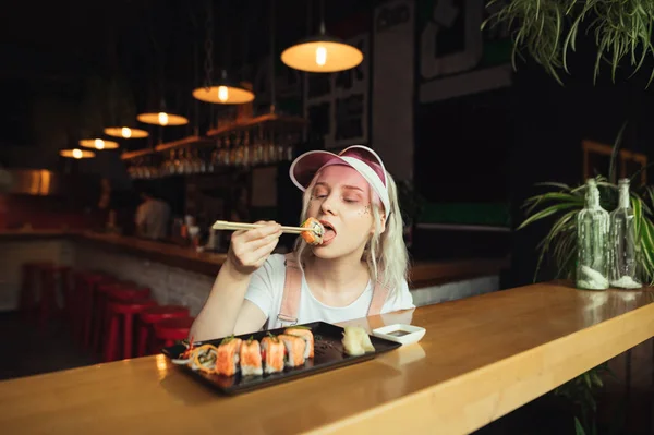 Süßes Mädchen mit Make-up und rosa Kappe isst Sushi mit Stäbchen in einem gemütlichen asiatischen Restaurant. Frau beißt genüsslich Sushi, am Wochenende isst sie gerne Sushi. Konzept der japanischen Küche. — Stockfoto