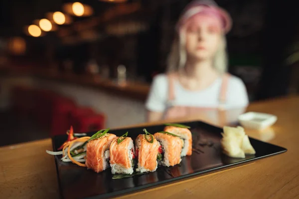 Тарелка с суши-роллами Филадельфии на столе в уютном ресторане, на фоне девушки в милой одежде. Аппетитные суши и девушка сидит за столом. Японская еда фото . — стоковое фото