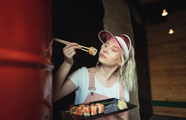 Model zjada Sushi Roll w restauracji, trzyma talerz i pałeczki. Atrakcyjna blondynka w różowej czapce stwarza na kamerę z rolką Sushi, portret w pomieszczeniu. Koncepcja japońskiego jedzenia. — Zdjęcie stockowe