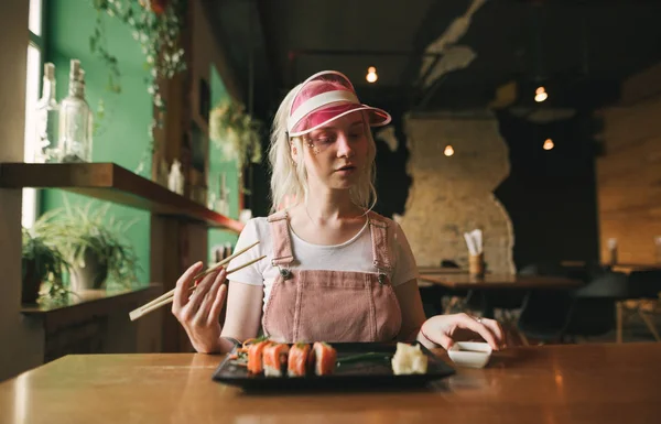 Attraktive Mädchen sitzt in einem gemütlichen japanischen Restaurant mit einem Teller Sushi-Brötchen und Sojasauce, hält Essstäbchen, schaut nach unten, trägt eine rosa Mütze und zieht sich aus. asiatisches Restaurantkonzept. — Stockfoto