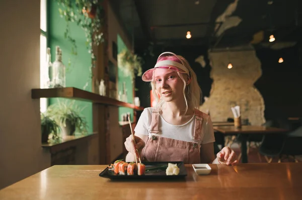 Lächelndes nettes Mädchen in pinkfarbener Kleidung, das in einem gemütlichen Restaurant Sushi-Rollen isst und zufrieden in die Kamera vor der Kulisse eines japanischen Restaurants blickt. Stilvolles Mädchen beim Mittagessen in der Sushi-Bar. — Stockfoto