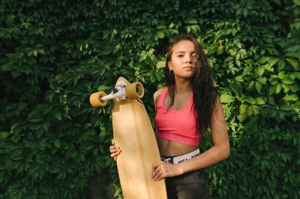 Closeup portrait of a Hispanic girl standing with a longboard in her hands against a background of green wall with ivy, looking into the camera with a serious face, wearing stylish street clothes — Stock Photo, Image