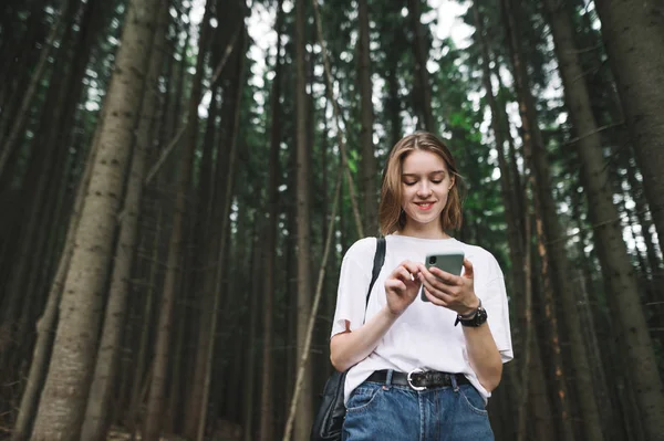 Sonriente joven turista usando smartphone en el bosque montañoso de coníferas. Viajadora alegre con amplia sonrisa enviando mensajes de texto a alguien a través de su teléfono sintiéndose feliz en el bosque de abetos. Copiar espacio . Imagen De Stock