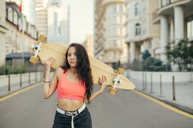 Uzun panolu çekici bir kızın sokak portresi, sokak manzarasının arka planında kameraya poz veriyor, kameraya bakıyor, şık günlük kıyafetler giyiyor. Longboard konsepti.