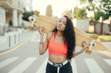 Elinde kaykayla gezen kızın sokak fotoğrafı. Turuncu tişörtlü çekici bir kızın portresi yolda ve gün batımında gözleri kapalı, elinde uzun panoyla caddede duruyor..