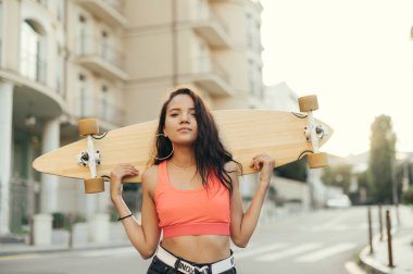 Pembe tişörtlü gülümseyen İspanyol kız elinde tahta ile kameraya bakıyor. Gün batımında elinde kaykayla gezen mutlu sokak binicisinin portresi..