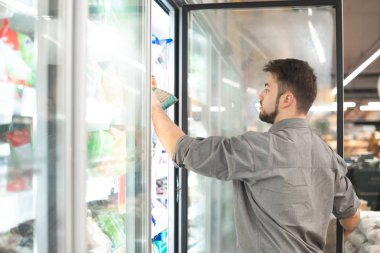 Man in his shirt takes his hand frozen foods from the refrigerator in the supermarket. The buyer selects the products in the grocery store's refrigerator. clipart