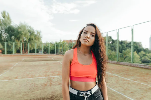 Retrato de cerca de una atractiva chica hispana con una camiseta naranja posando frente a la cámara sobre el fondo de una vieja pista de tenis. Concepto de calle . — Foto de Stock