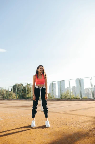 Foto de menina elegante feliz em roupas casuais em pé no parque infantil, olhando para longe e sorrindo. Menina hispânica com cabelos encaracolados escuros posa na câmera e sorri, fundo de uma antiga quadra de tênis . — Fotografia de Stock