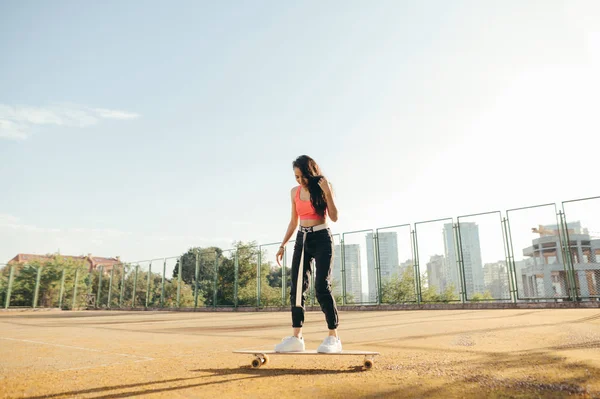 Elegante ragazza in abiti da strada cavalca su un longboard sul parco giochi. Attraente ragazza ispanica in abiti casual si erge su un pattino, foto di strada, sfondo di un vecchio campo da tennis. Concetto Longboard . — Foto Stock