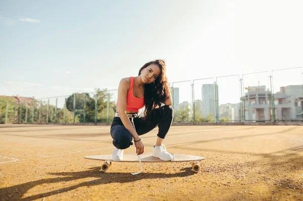 Chica hispana de moda sentada en un longboard y posando para la cámara, usando ropa de calle con estilo y zapatillas blancas. —  Fotos de Stock