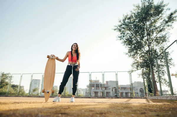 Chica hispana feliz de pie con longboard en el patio de recreo y sonriendo, con ropa de calle con estilo. Chica atractiva con un monopatín posando en la cámara en el fondo del atardecer . —  Fotos de Stock