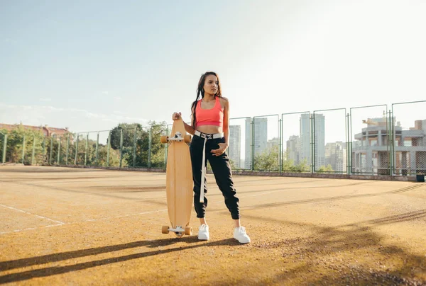 Menina atraente em roupas de rua na moda fica com um longboard na mão no parque infantil, posando para a câmera, olhando para longe. Modelo menina hispânica com skate em mãos fica no velho campo de ténis . — Fotografia de Stock