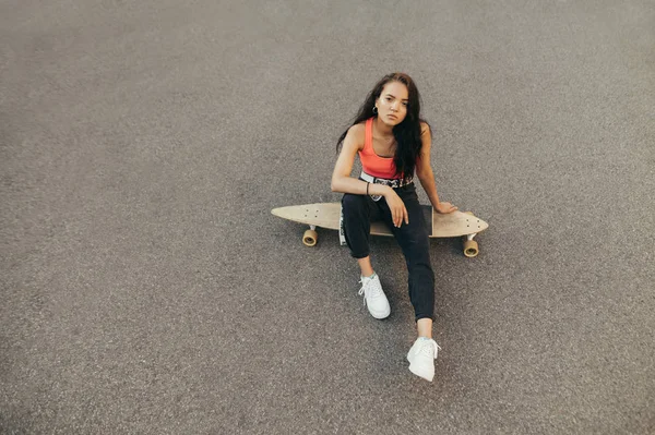 Portrait d'une jolie fille assise sur asphalte avec longboard, regardant la caméra avec un visage sérieux, portant des vêtements décontractés élégants. Fille hispanique avec planche à roulettes isolé sur fond d'asphalte — Photo