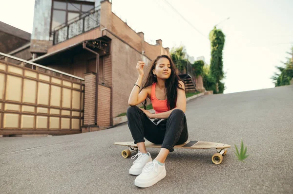 Rapariga mulata sorridente sentada no logboard no fundo da rua, olhando para baixo, vestindo roupas de rua. Menina patinadora relaxante em longboards em asfalto, positivo, cultura de rua . — Fotografia de Stock