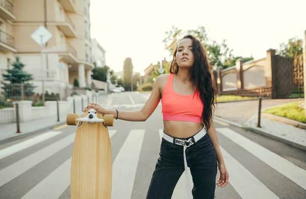 Attractive girl in a pink T-shirt and jeans stands on the street with a longboard in her hand on a street background at sunset. Hispanic girl posing with a logboard on the street in the summer evening