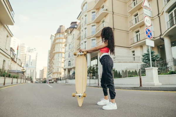 Ragazza alla moda cavaliere con skateboard all'aperto la sera calda. Street sera ritratto di una donna in piedi lateralmente con un longboard in mano, guardando altrove, su uno sfondo di strada — Foto Stock